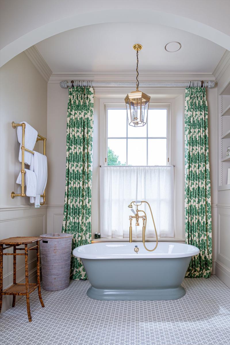 Freestanding bathtub with brass taps beneath an arched window with green botanical curtains